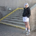 Kobieta w sportowej stylizacji – biała bluza z kapturem, czarna pikowana kurtka, białe szorty, długie białe skarpetki Basic Arctic Snow i szare buty trekkingowe – stoi przy betonowych schodach z żółtą krawędzią.