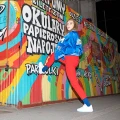 Kobieta w czerwonych spodniach i białych sneakersach pozuje na tle kolorowego graffiti, eksponując jeansowe skarpetki Basic w stylu streetwear.