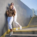 Dwie kobiety w jasnych stylizacjach na betonowych schodach. Jedna w beżowych skarpetkach Peach Beach, druga w brązowych. Gładkie, bawełniane skarpetki dodają stylizacji lekkości i miejskiego luzu.