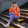 Mężczyzna w jasnożółtych skarpetkach Basic - Banana Shake, pomarańczowej bluzie z kapturem i fioletowych spodniach, siedzący na drewnianych schodach w kolorowych sneakersach.