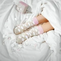 Kobieta leży pod białą kołdrą w łóżku, ma na sobie beżowe skarpety z różowym ściągaczem i wzorem słodkich pianek marshmallow – wokół niej rozsypane są pianki, obok leży pudełko „Marshmallows