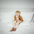 Uśmiechnięty chłopiec w brązowej koszulce siedzi na łóżku w białej pościeli, trzyma puszkę "Hot Chocolate" i je bitą śmietanę. Na stopach ma dziecięce skarpetki.