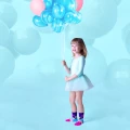 Dziewczynka w jasnoniebieskiej sukience baletowej trzyma bukiet różowo-niebieskich balonów, stojąc na tle dużych pastelowych balonów, ma na sobie kolorowe skarpetki z bajkowymi wzorami.