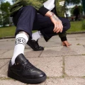 Mężczyzna w czarnych butach i białych skarpetkach z dużym napisem "NINETIES” i uśmiechniętą buźką, przyklęknięty na miejskim chodniku..
