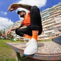 Mężczyzna w pomarańczowej koszulce i czapce z daszkiem kuca na betonowym stole w blokowisku. Na nogach ma białe sneakersy i pomarańczowe skarpetki z napisem  „NINETIES”  i uśmiechem.