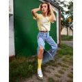 Kobieta w żółtych skarpetkach z białym napisem  „NINETIES”, żółtym T-shircie i jeansach z rozcięciami, pozująca dynamicznie na tle zielonej bramy.
