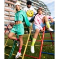 Trzy osoby w kolorowych dresach i skarpetkach „NINETIES”. Stoją i siedzą na metalowym drabinkowym placu zabaw, na tle blokowiska.