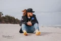 Mężczyzna siedzący na piaszczystej plaży w jeansach i czarnej kurtce, ma na stopach żółte skarpetki we wzór pizzy..