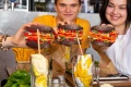 Troje osób w restauracji trzyma w dłoniach kolorowe skarpetki ułożone w kształt burgerów nad stołem z napojami i przekąskami.
