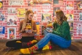 Dwie kobiety siedzące na podłodze w retro wnętrzu, trzymające opakowania w kształcie burgerów z kolorowymi skarpetkami oraz mające na stopach skarpetki w burgerowy wzór.