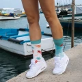 Kolorowe skarpetki z motywem morskich stworzeń w pastelowych barwach, założone do białych sneakersów; modelka stoi na nabrzeżu przy łodziach.