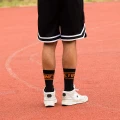 Mężczyzna w sportowych butach i czarnych skarpetkach z napisem „NINETIES” w pomarańczowym kolorze, stojący na tartanowym boisku w czarno-białych szortach.