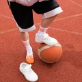 Dziecko w białych skarpetkach z pomarańczowymi płomieniami i napisem „NINETIES”. Stawia nogę na pomarańczowej piłce do koszykówki na boisku. Energiczna, sportowa stylizacja z ogniem w kroku.