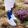Stopy w białych sneakersach i białych spodniach, ubrane w kolorowe skarpetki mixTURY Pink Flamingo w odcieniach niebieskiego i różu, przedstawiające flamingi; osoba stoi na kamienistej ścieżce obok kęp fioletowych kwiatów.