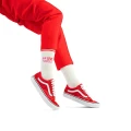 Modelka w czerwonych spodniach i czerwonych sneakersach prezentuje białe skarpetki za kostkę z napisem „LOVE YOURSELF” w kolorze czerwonym i różowym oraz z czerwonym paskiem na ściągaczu. Tło białe.