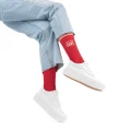 Osoba w jasnych dżinsach i białych sneakersach siedząca z uniesionymi nogami, na stopach czerwone skarpetki z napisem „BE MY LOVE” na białym tle.