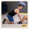 Mężczyzna w sportowym stroju klęczy na jasnej podłodze, trzymając nogi partnerki wykonującej brzuszki; oboje mają na sobie kolorowe, czarne skarpetki z antypoślizgowym spodem i wzorem w różowym, żółtym i niebieskim kolorze.
