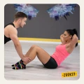 Para trenuje razem w sali fitness – kobieta w różowym topie i czarnych szortach wykonuje brzuszki, mężczyzna w sportowym stroju trzyma jej stopy. Oboje mają na sobie czarne, kolorowe skarpetki z antypoślizgowym wzorem, idealne na trening.