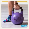 Zbliżenie na nogę w sportowej, fioletowej skarpetce z antypoślizgowym wzorem ABS i kolorowym printem, obok ręki trzymającej 20-kilogramowy odważnik kettlebell LEON na drewnianej podłodze w siłowni.