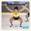 Kobieta w pozycji przysiadu z uniesioną sztangą, ubrana w sportowy top i antypoślizgowe skarpetki treningowe w kolorach granatu, żółci i błękitu, na tle jasnej sali fitness z kolorową grafiką skrzydeł na ścianie.