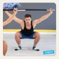 Mężczyzna w przysiadzie z uniesioną sztangą, ubrany w czarną koszulkę, niebieskie spodenki i antypoślizgowe skarpetki sportowe w kolorach granatu, błękitu i żółci, ćwiczy na jasnej podłodze w sali fitness z szarą ścianą