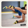 Dwie osoby ćwiczące na sali fitness w kolorowych, antypoślizgowych skarpetkach sportowych z silikonowymi wypustkami na spodzie; dynamiczna poza z uniesionymi nogami, widoczna drewniana podłoga i szare ściany.