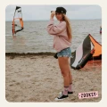 Kobieta w różowej bluzie, jeansowych szortach i różowych skarpetkach z napisem ‘PARADISE’ stoi na plaży przy wodzie, w tle widoczne są skrzydło do kitesurfingu i osoba na desce