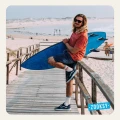 Uśmiechnięty mężczyzna z długimi blond włosami i okularami przeciwsłonecznymi siedzi na barierce drewnianego pomostu z deską surfingową. Ma na sobie czerwony T-shirt, dżinsowe szorty, sportowe buty i kremowe skarpetki z napisem SUNSHINE i wzorem słońca
