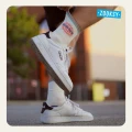 Mężczyzna w białych sneakersach i białych skarpetkach z kolorowym wzorem słońca oraz napisem SUNSHINE poprawia ściągacz jednej ze skarpet. Tło to miejska przestrzeń z rozmytym budynkiem i drzewem.