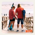 Para stojąca tyłem na drewnianym pomoście przy plaży, ubrana w kolorowe skarpetki z napisem ALOHA i SUNSHINE, z widokiem na ocean w tle