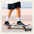 Dziewczyna jadąca na deskorolce w białych skarpetkach z ribowaną cholewką i granatową grafiką fali, ubrany w granatowe szorty i czarne sneakersy, na tle piaszczystej plaży i oceanu.