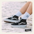 Zbliżenie na nogi dziewczyny w białych skarpetkach z ribowaną cholewką i granatowym wzorem fali, ubrane w czarne sneakersy Vans; dziewczyna siedzi na jasnym brukowanym podłożu