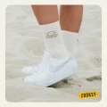 Nogi w białych sneakersach stojące na piasku, ubrane w jasne skarpetki z ribowaną cholewką i brązowym wzorem zachodzącego słońca na górze cholewki