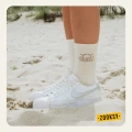 Kobiece nogi w białych sneakersach stojące na piasku, w jasnych skarpetkach z ribowaną cholewką i brązowym wzorem zachodzącego słońca z falami i sercami na górze cholewki