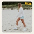Kobieta spacerująca po piaszczystej plaży w białej bluzie, dżinsowych szortach i jasnych skarpetkach z ribowaną cholewką, ozdobionych brązowym wzorem zachodzącego słońca na górze cholewki