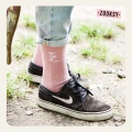 Osoba stojąca na ziemi w czarnych sneakersach i różowych skarpetkach z ribowaną cholewką, ozdobionych białym wzorem dwóch palm na górze cholewki, z podwiniętymi dżinsami