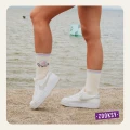 Zbliżenie na nogi kobiety stojącej przy brzegu wody; ma na sobie białe sneakersy i kremowe skarpetki z napisem ‘POWER’ oraz motywem yin-yang. W tle widoczne fale i pływające boje