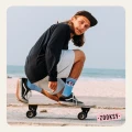 Młody mężczyzna w czapce z daszkiem jadący na deskorolce w pozycji kucznej, na tle plaży i morza, w błękitnych skarpetkach z różowym wzorem oka i napisem ‘ON MY MIND