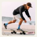 Skateboarder w czarnej bluzie, jasnych szortach i błękitnych skarpetkach z wzorem oka i napisem ‘ON MY MIND’ odbija się nogą podczas jazdy na deskorolce przy plaży