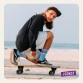 Młody mężczyzna w czapce z daszkiem i czarnym longsleevie jadący na deskorolce w pozycji kucznej, na tle plaży i morza, w błękitnych skarpetkach z różowym wzorem oka i napisem ‘ON MY MIND