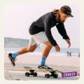 Skateboarder w czarnej bluzie, jasnych szortach i błękitnych skarpetkach z wzorem oka i napisem ‘ON MY MIND’ odbija się nogą podczas jazdy na deskorolce przy plaży