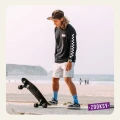 Młody mężczyzna w błękitnych skarpetkach z motywem oka i napisem 'ON MY MIND' stoi z longboardem na nadmorskim deptaku. W tle plaża, morze i klify
