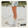 Kobieta w białych skarpetkach z brązowym wzorem słońca, jasnych sneakersach i dżinsowych szortach stoi na piaszczystej plaży