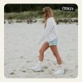 Kobieta w białych skarpetkach z motywem zachodzącego słońca spaceruje po plaży w białej bluzie, dżinsowych szortach i białych sneakersach