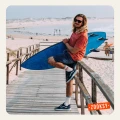 Uśmiechnięty mężczyzna z długimi blond włosami i okularami przeciwsłonecznymi siedzi na barierce drewnianego pomostu z deską surfingową. Ma na sobie czerwony T-shirt, dżinsowe szorty, sportowe buty i kremowe skarpetki z napisem SUNSHINE i wzorem słońca.