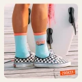 Skarpetki w kolorze miętowym z napisem „ALOHA” w stylu retro, widoczne na nogach osoby stojącej na drewnianym pomoście. Skarpetki mają pomarańczowo-różowy ściągacz, a napis otoczony jest różowymi promieniami i niebieską falą.