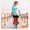 Kobieta w turkusowej koszulce tie-dye i czarnych kolarkach stoi na drewnianym pomoście z deską surfingową, ma na sobie błękitne skarpetki z letnim motywem i tenisówki w kratę.
