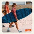 Para surferów idzie po deptaku przy plaży; kobieta niesie deskę surfingową w niebieskim pokrowcu i ma na sobie błękitne skarpetki z pomarańczowym motywem i czarno-białe tenisówki w kratkę