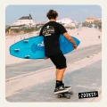 Mężczyzna w czarnym zestawie ubraniowym jedzie na deskorolce po deptaku przy plaży, trzymając niebieską deskę surfingową pod pachą. Na sobie ma czarne skarpetki z białym wzorem deski surfingowej i czarne trampki. W tle zabudowania nadmorskiego miasteczka