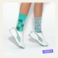 Dziecięce nogi w białych sneakersach, kolorowych świątecznych skarpetkach. Po lewej morska skarpetka w wieńce i kule śnieżne, po prawej szara z prezentami, gałązkami i cukrowymi laskami.